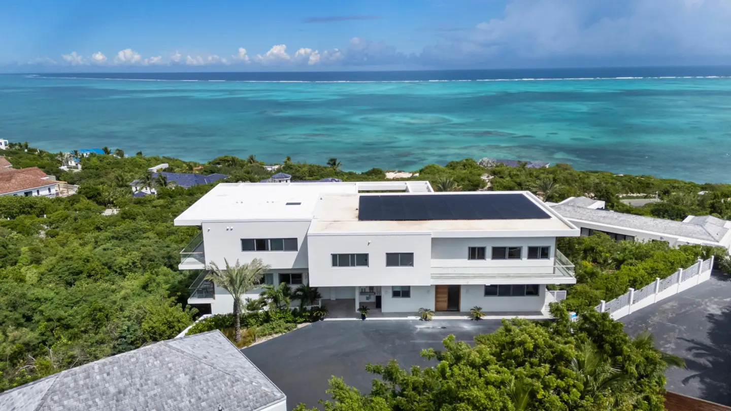 Aerial view of a modern white house with solar panels, surrounded by lush greenery, overlooking a turquoise ocean.