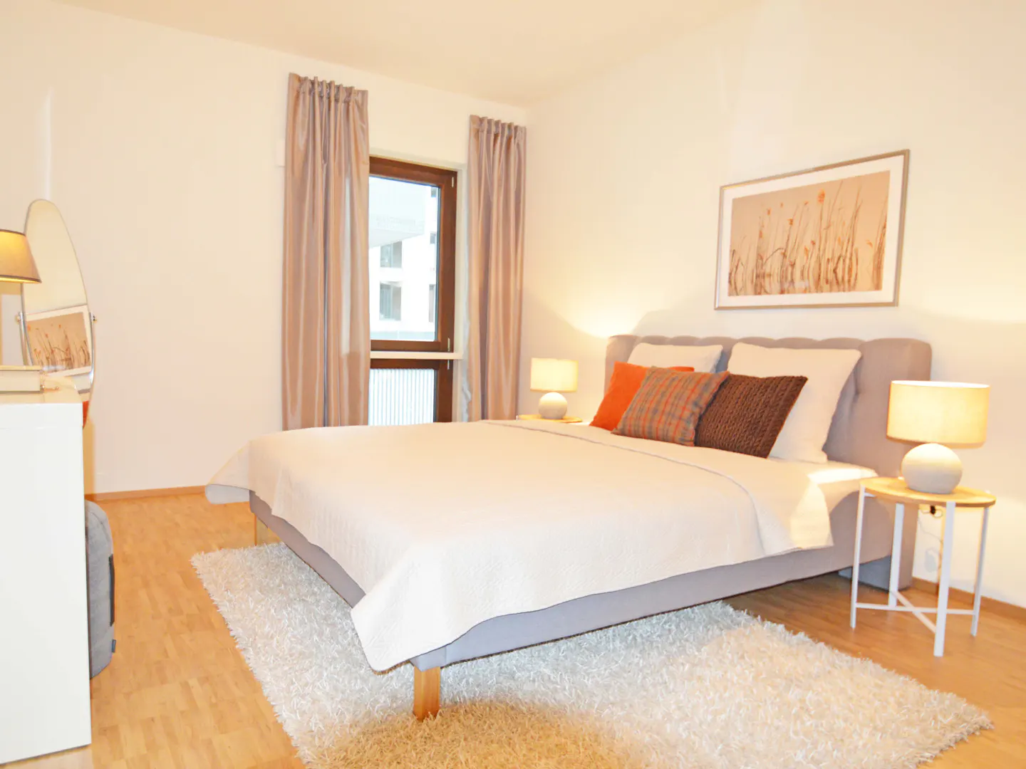 Bedroom with a gray bed, white bedding, and orange and brown pillows. Two bedside tables with lamps, a window with curtains, and a picture on the wall.