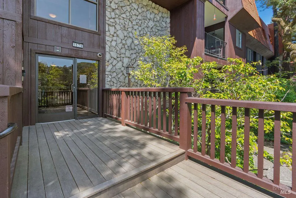 Exterior view of a brown building with a stone facade and a wooden deck. Glass doors lead into the building. Green trees are visible in the background.
