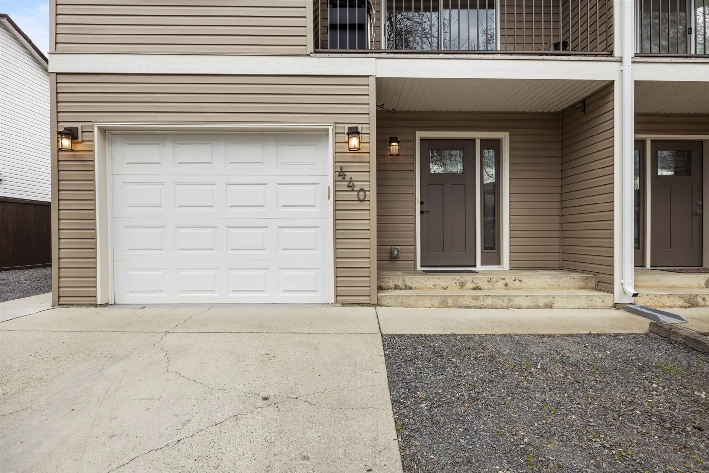 Exterior view of a two-story townhouse with beige siding, a white garage door, and a brown front door. Balconies are visible on the second floor.