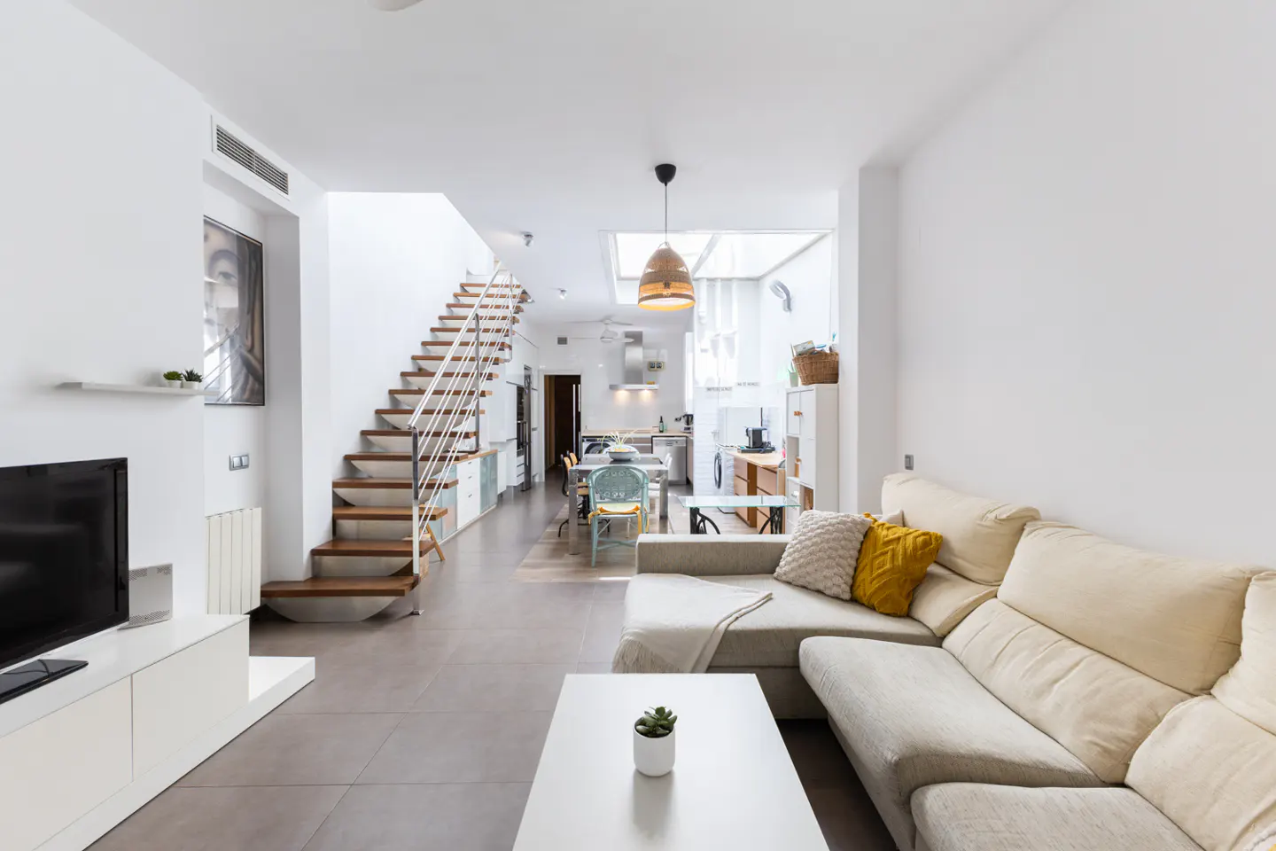 Modern open-plan living room with a beige sectional sofa, white coffee table, wooden staircase, and kitchen in the back.