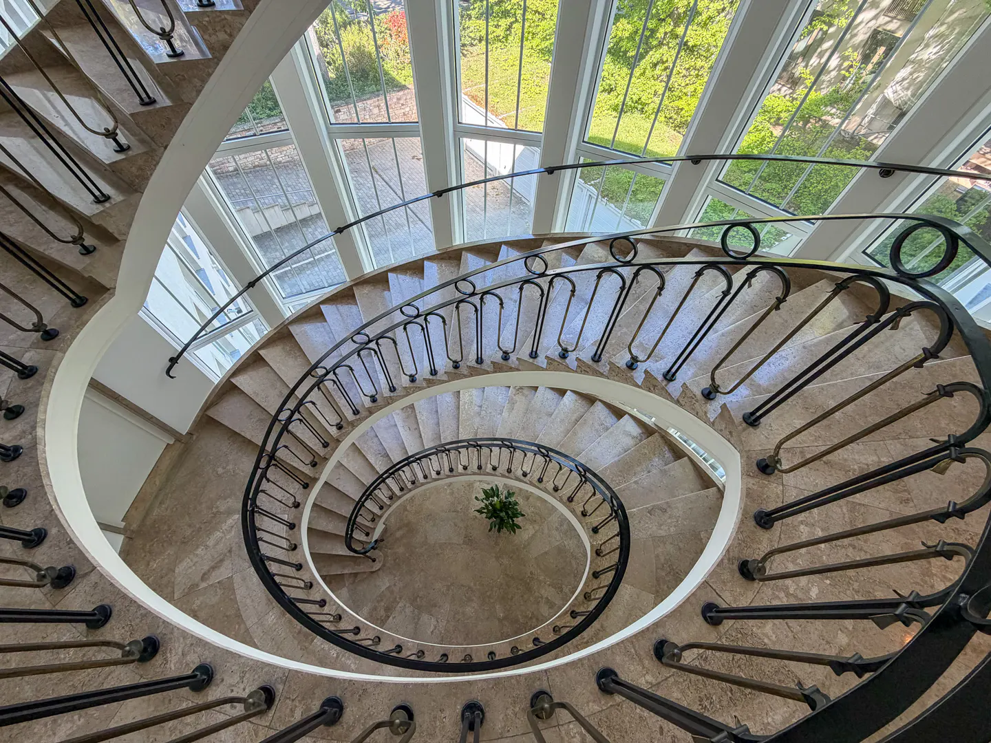 A high-angle shot of a spiral staircase with beige marble steps and a black metal railing. A small green plant sits at the bottom. Windows are visible above.