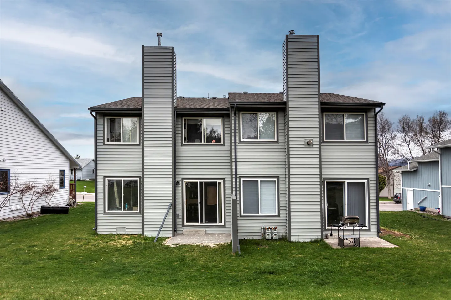 Back of a two-story gray townhouse with two chimneys, windows, and sliding glass doors, on a green lawn.