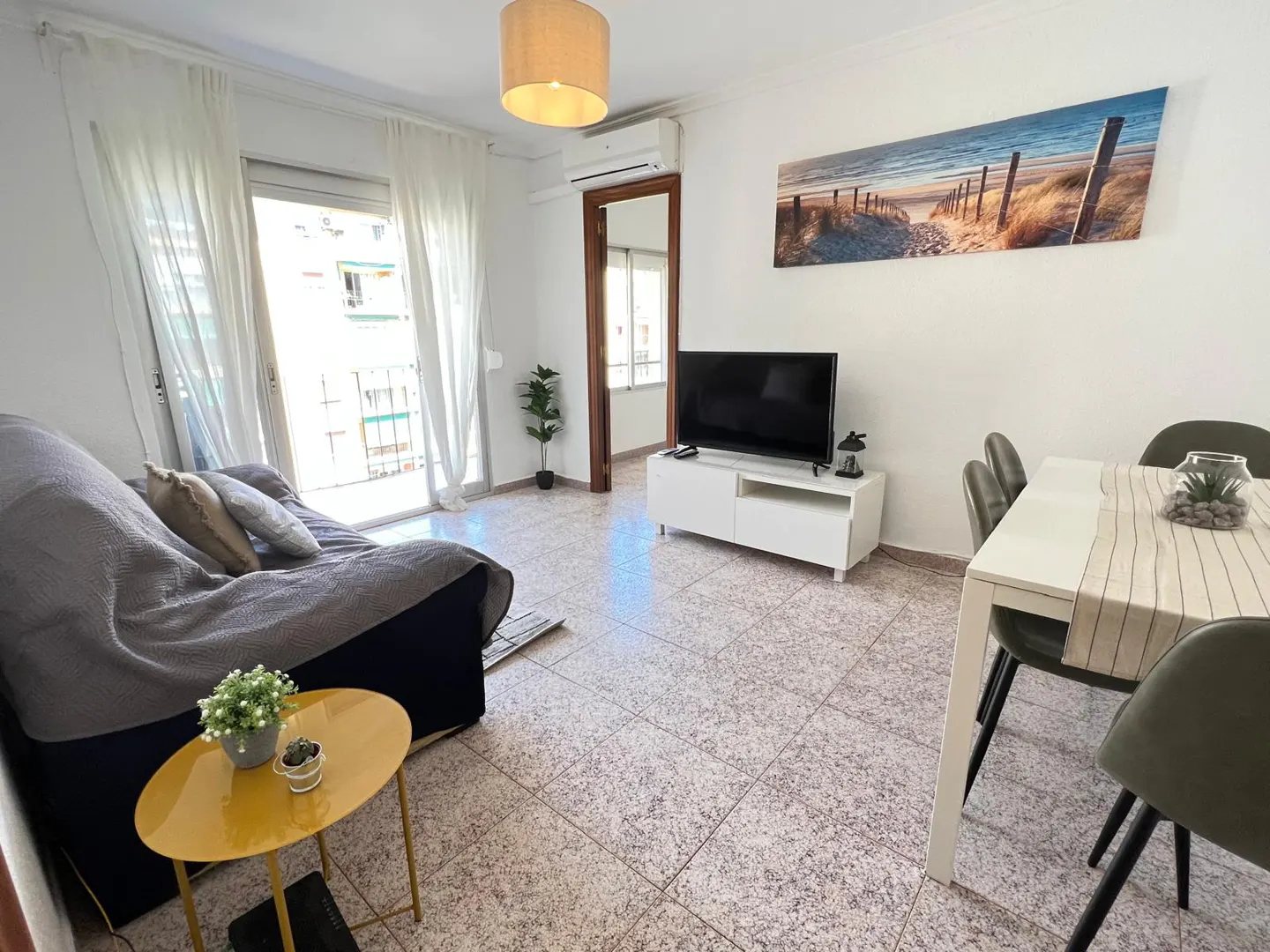 Bright living room with a grey sofa, yellow coffee table, white TV stand, and a dining area with a beach wall art piece.