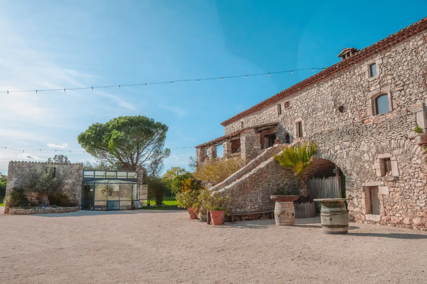 Exterior of a stone building with a staircase, potted plants, and a greenhouse under a blue sky.