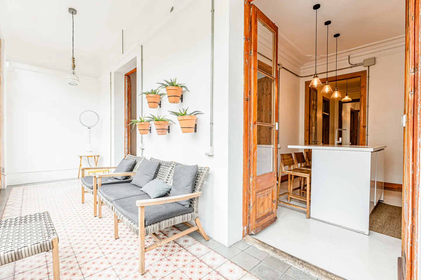 Bright, airy room with seating area, potted plants, and open doorway to a kitchen with a white island and pendant lights.