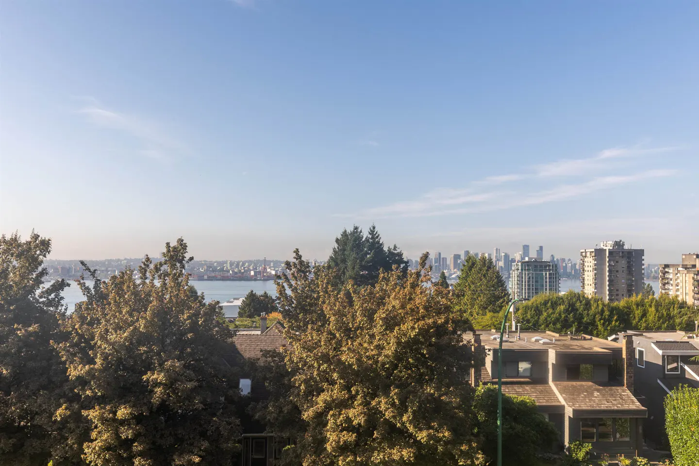 View of Vancouver skyline and harbor from a residential neighborhood. Trees and rooftops in the foreground. Blue sky.
