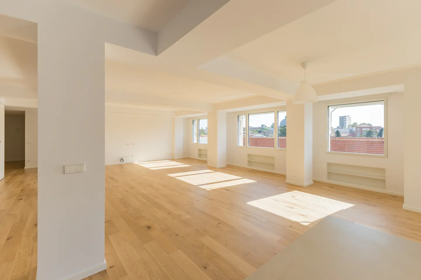 Bright, empty room with light wood floors, white walls, and large windows. Sunlight streams in, illuminating the space.