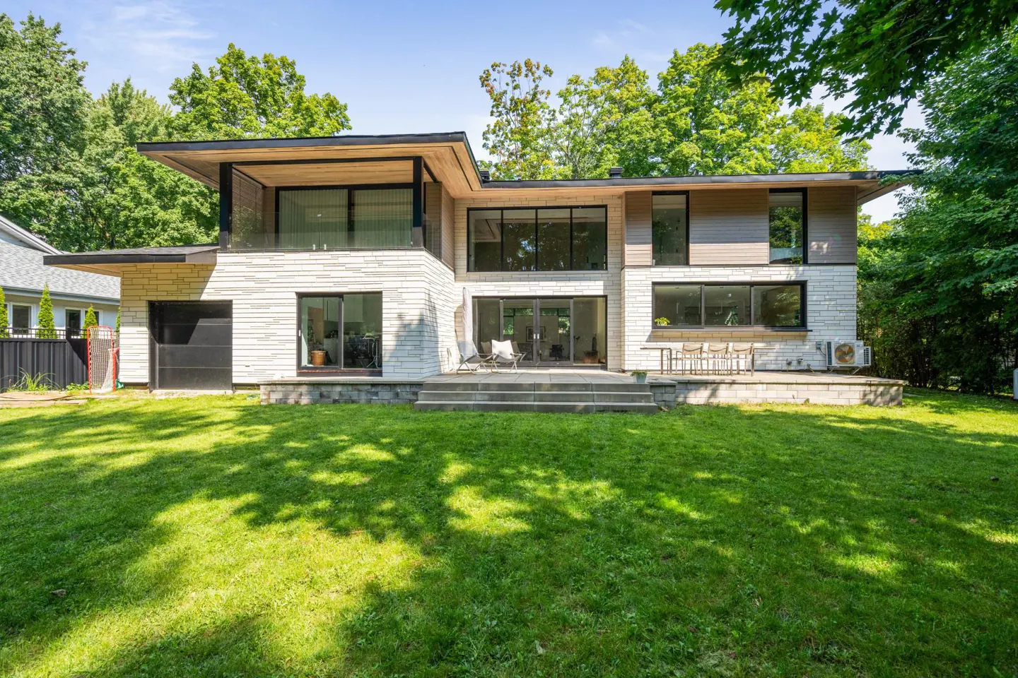 Two-story modern home with stone and wood siding, black trim, and large windows. Green lawn in front, trees in background.