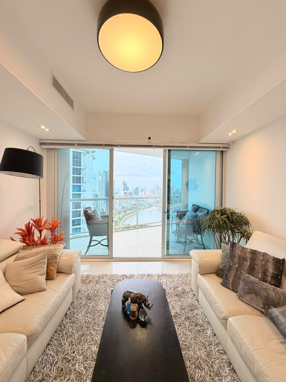 Modern living room with two white sofas, a black coffee table, and a balcony overlooking a city skyline and water. [AI-generated text]