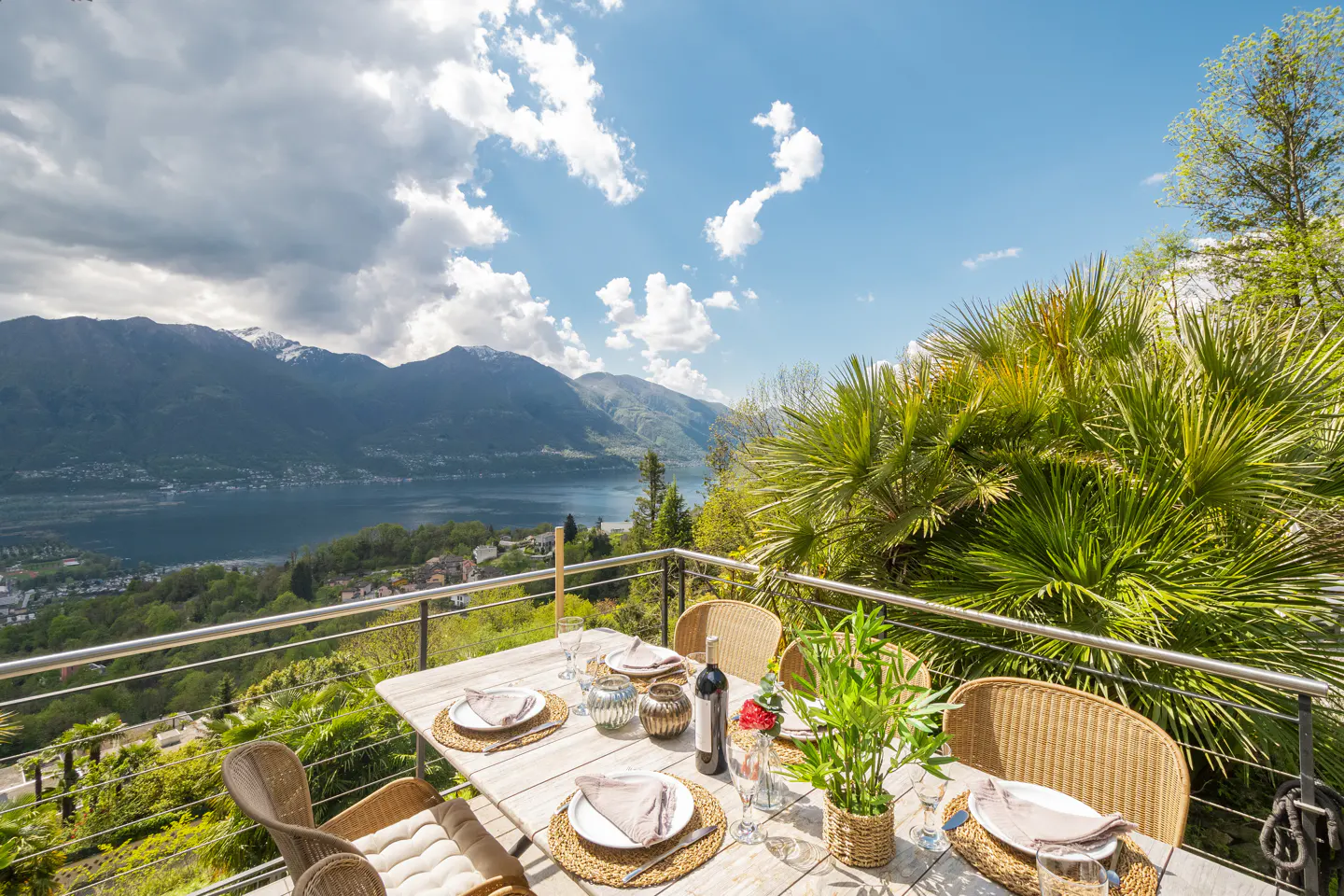Outdoor dining table set with plates, wine, and flowers on a balcony overlooking a lake and mountains.