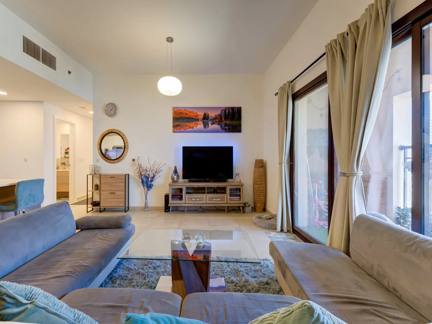 Living room with gray sofas, glass table, and TV. Beige curtains frame a sliding glass door to a balcony. Artwork and decor add color.