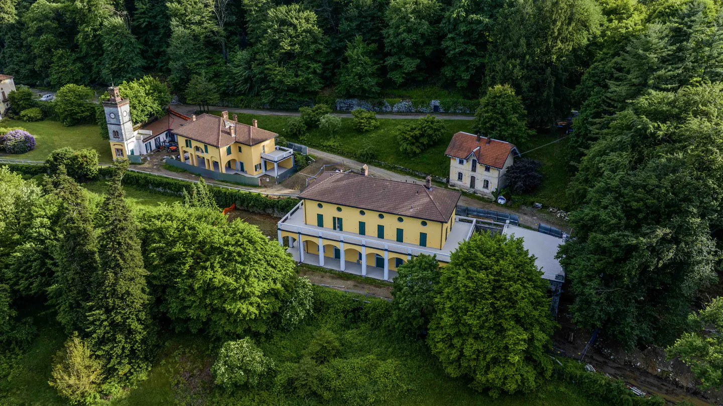Aerial view of yellow buildings with red roofs surrounded by lush green trees. A clock tower is visible on the left.