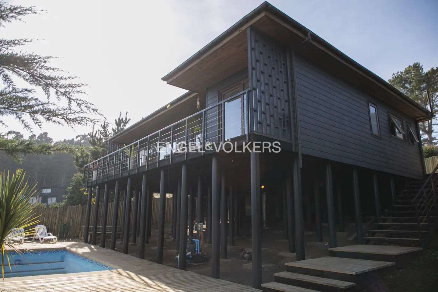 Modern dark-gray stilt house with a balcony, wooden deck, and swimming pool in a wooded setting.