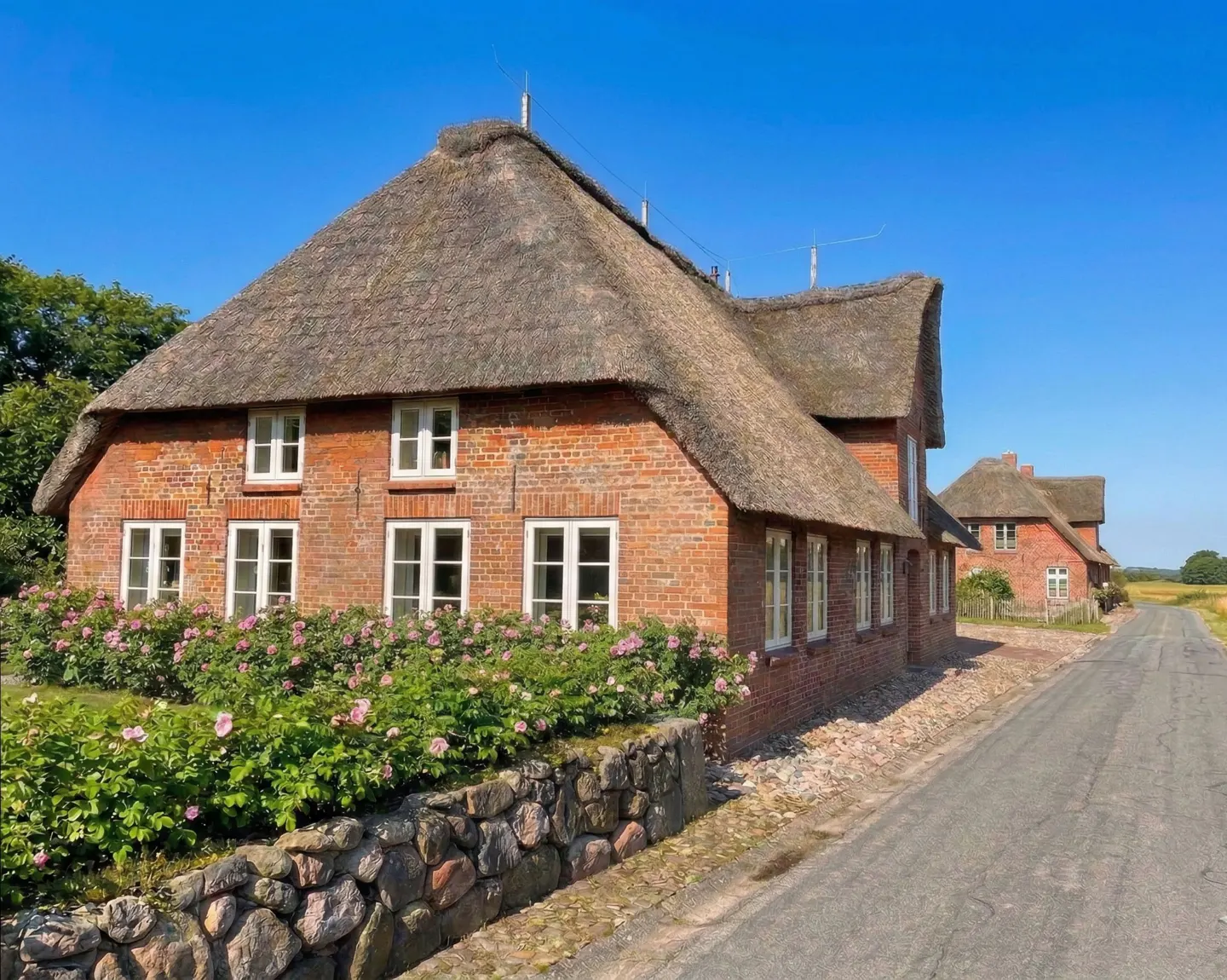 A red brick house with a thatched roof and white windows, with a stone wall and pink flowers.