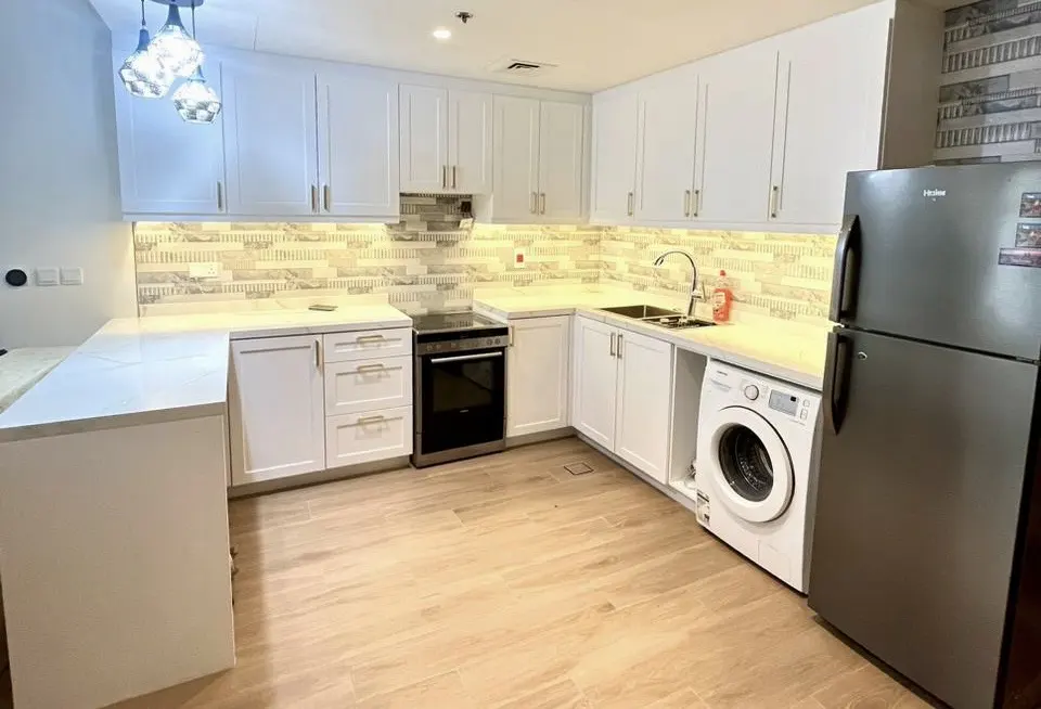 Bright kitchen with white cabinets, marble countertops, and wood-look tile flooring. Stainless steel refrigerator and washing machine are visible.