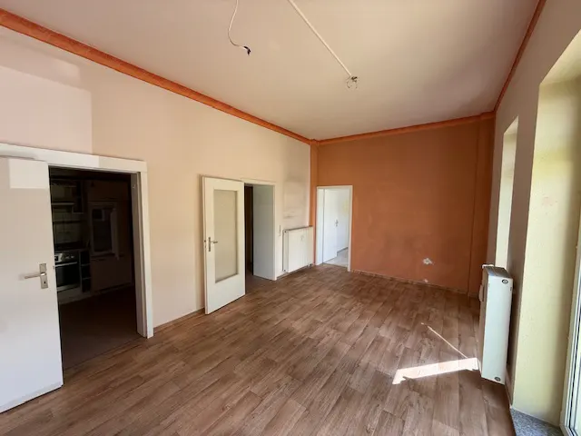 Empty room with wood floors, peach and orange walls, and white trim. Doors lead to other rooms, including a kitchen.