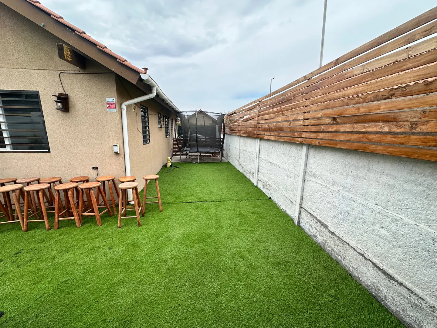 A backyard with green artificial turf, a row of wooden stools, and a trampoline in the background. A wooden fence tops a concrete wall.