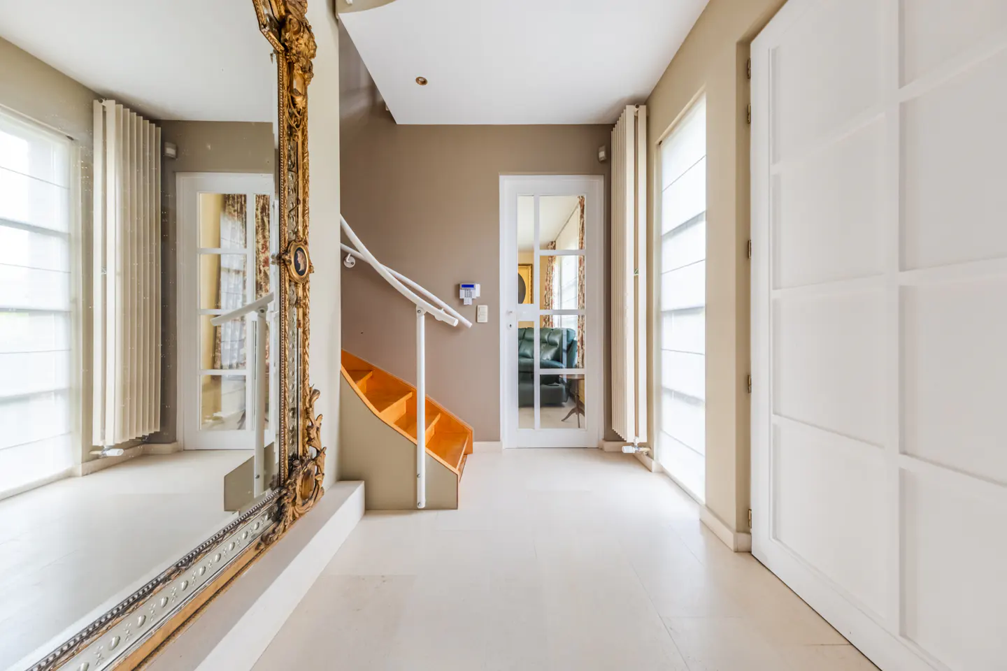 Bright hallway with a large ornate gold mirror, white paneled doors, and a small staircase with orange steps.