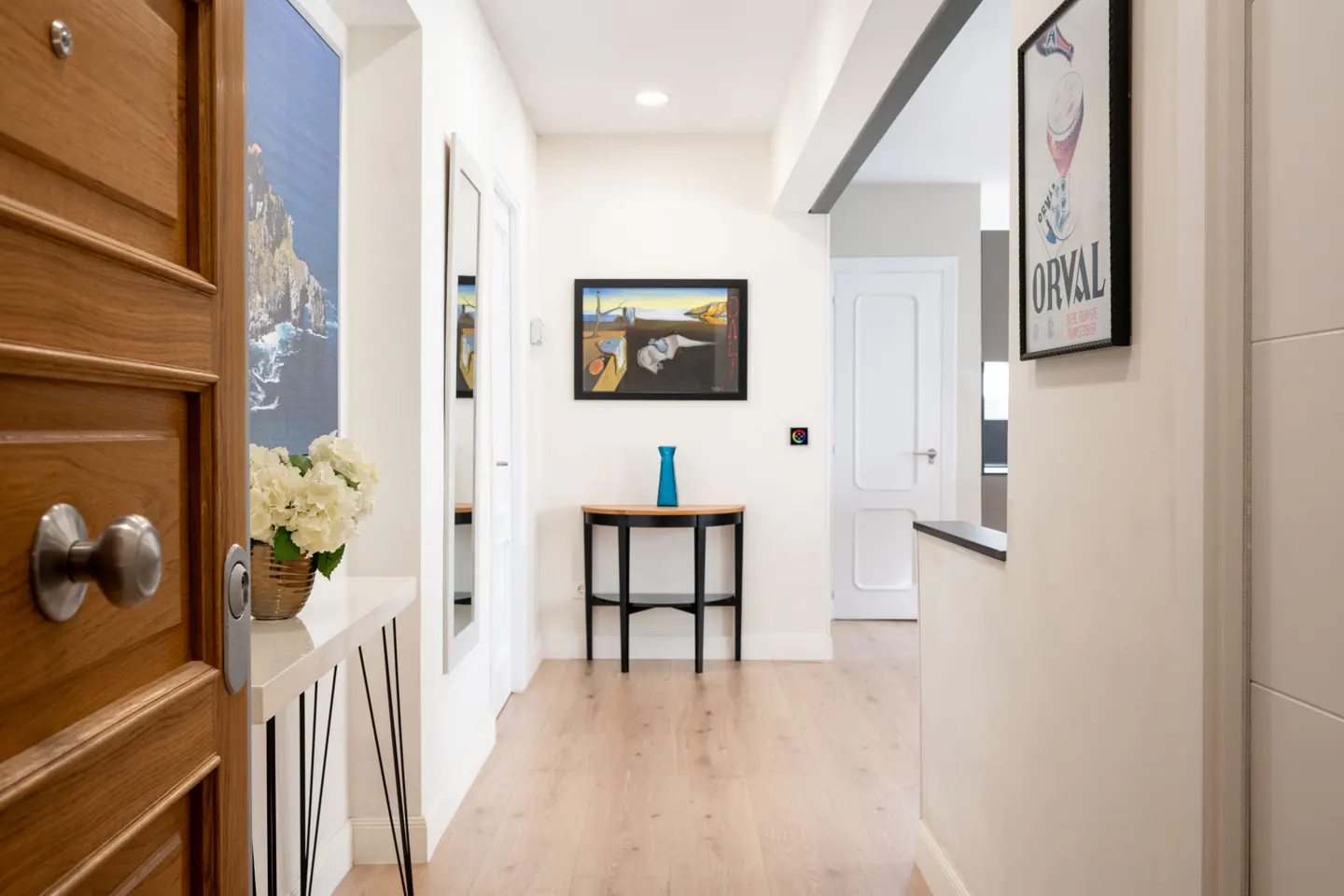 Open wooden front door reveals a bright hallway with light wood floors, white walls, and decorative art.