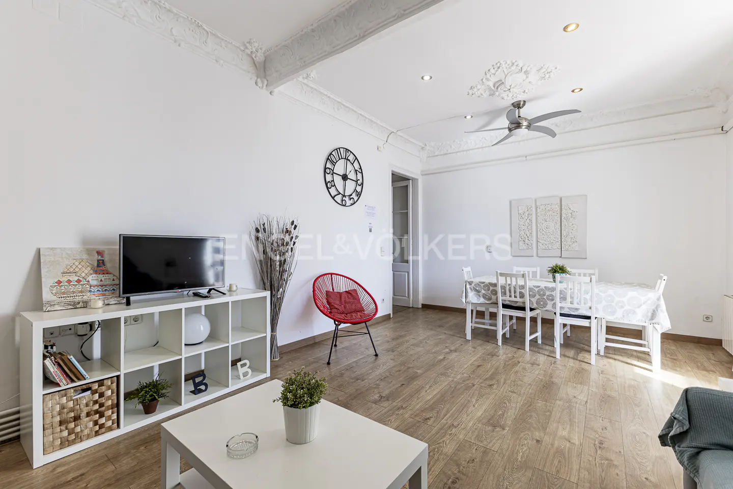 Bright living room with white walls, wood floors, and ornate ceiling. Features a TV, shelving unit, dining table, and red chair.
