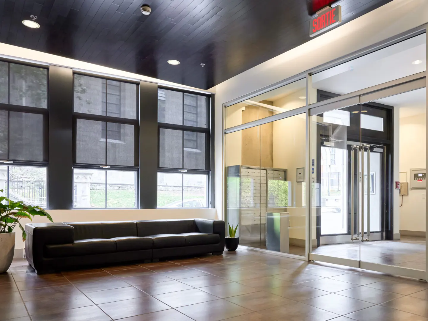 Condo lobby with a black leather sofa, large windows, and glass doors. The floor is brown tile, and there are plants in pots. An exit sign is visible.