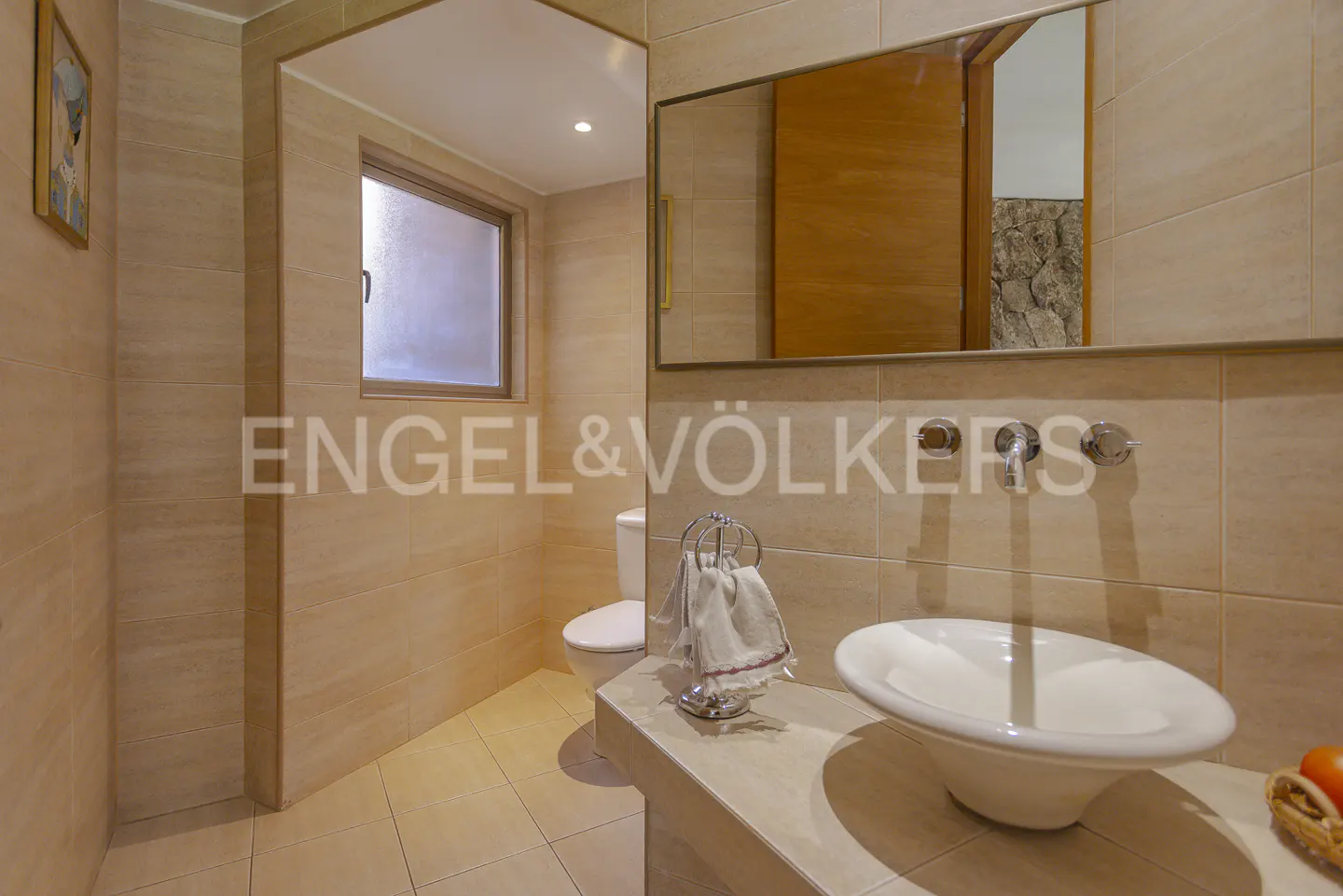 Bathroom with beige tile walls, a white sink, a toilet, and a mirror reflecting a wooden door and stone wall.
