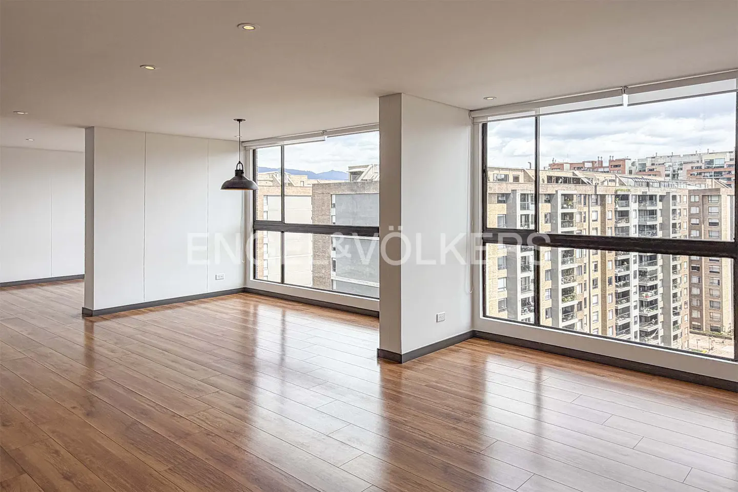 Bright, empty room with hardwood floors, white walls, and large windows overlooking a city skyline. A black pendant light hangs from the ceiling.