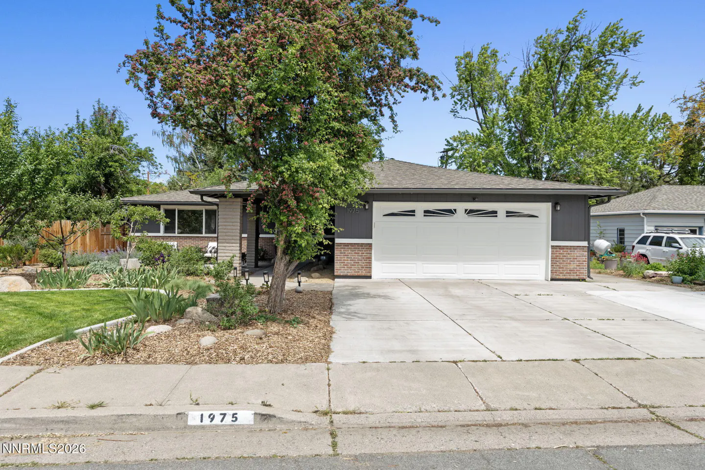 A single-story house with gray siding, a white garage door, and a well-manicured lawn. A tree is in the front yard.