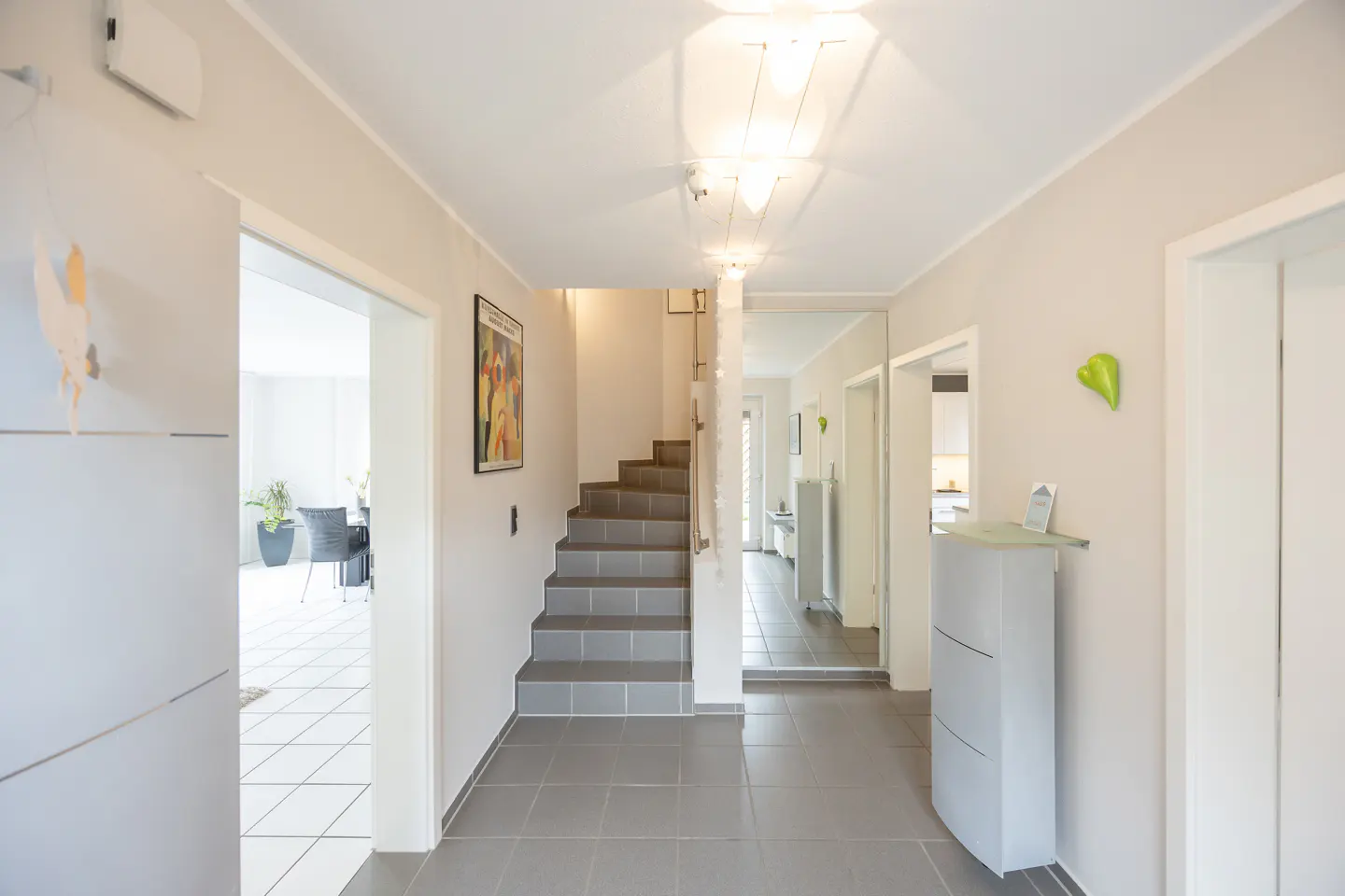 Bright hallway with gray tile floor, white walls, and a staircase. A mirror reflects the hallway, and a doorway leads to a dining area.