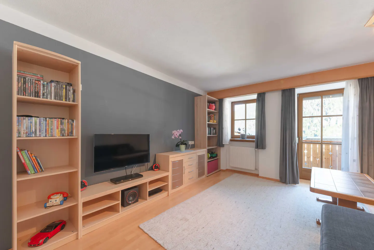 Living room with gray wall, light wood furniture, TV, bookshelf with DVDs, and a door to a balcony.