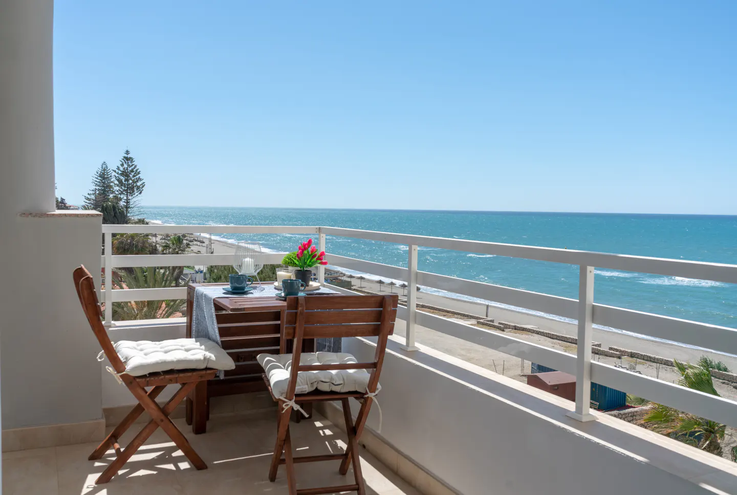 Balcony with a table, chairs, and ocean view. The table is set for two with flowers. The railing is white.