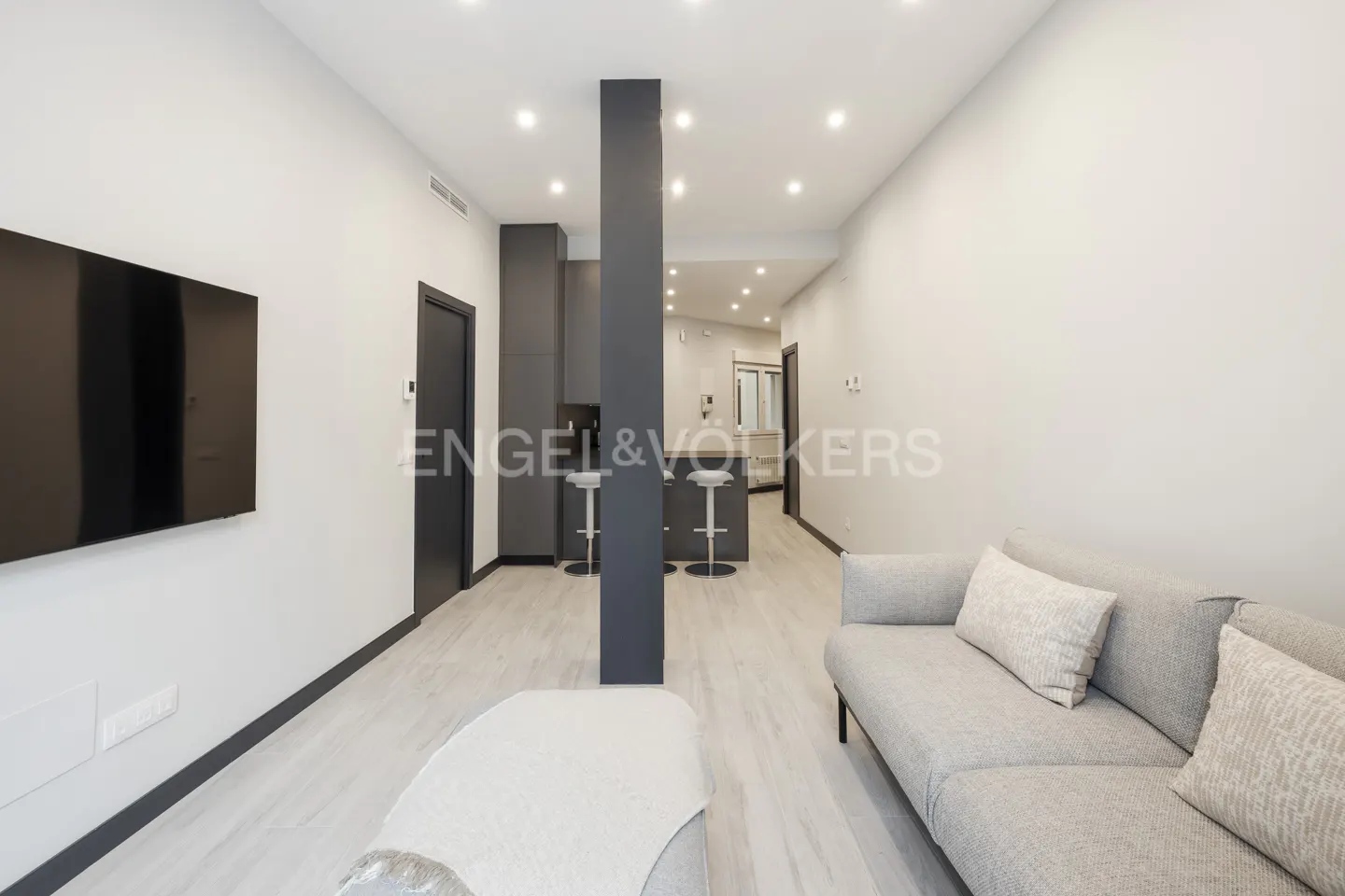 A modern apartment interior with a gray sofa, TV, and kitchen with bar stools. The walls are white, and the floor is light wood.