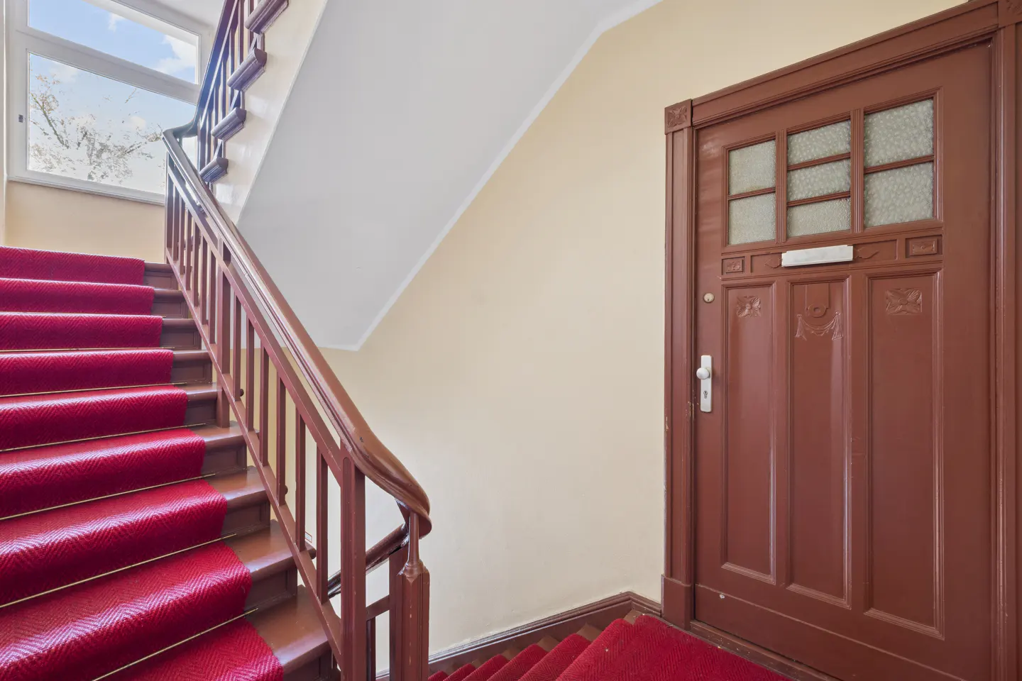 Interior shot of a staircase with a red carpet and a brown wooden door.
