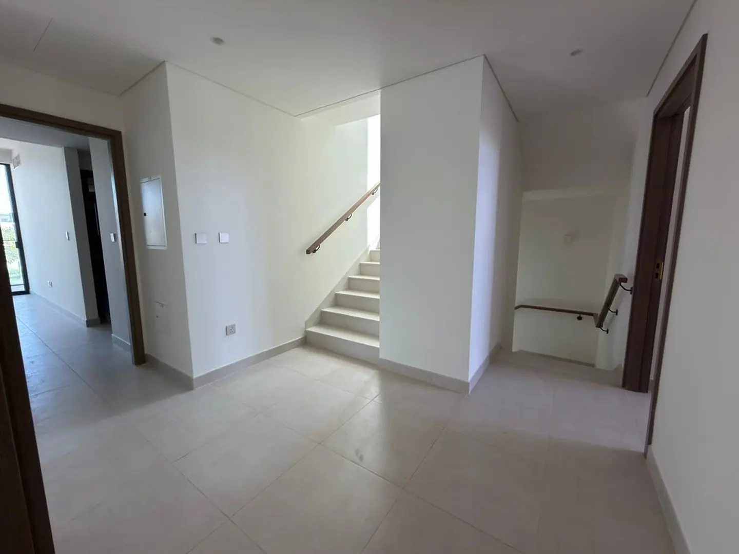 Modern hallway with white walls, light tile floors, and a staircase with a wooden handrail leading to an upper level.