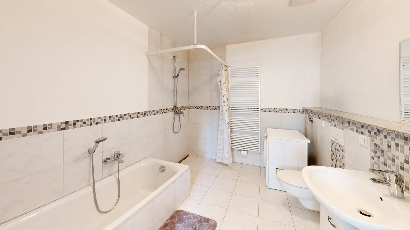 Bright bathroom with white tile, mosaic trim, and white fixtures. A tub with shower, toilet, sink, and towel rack are visible.