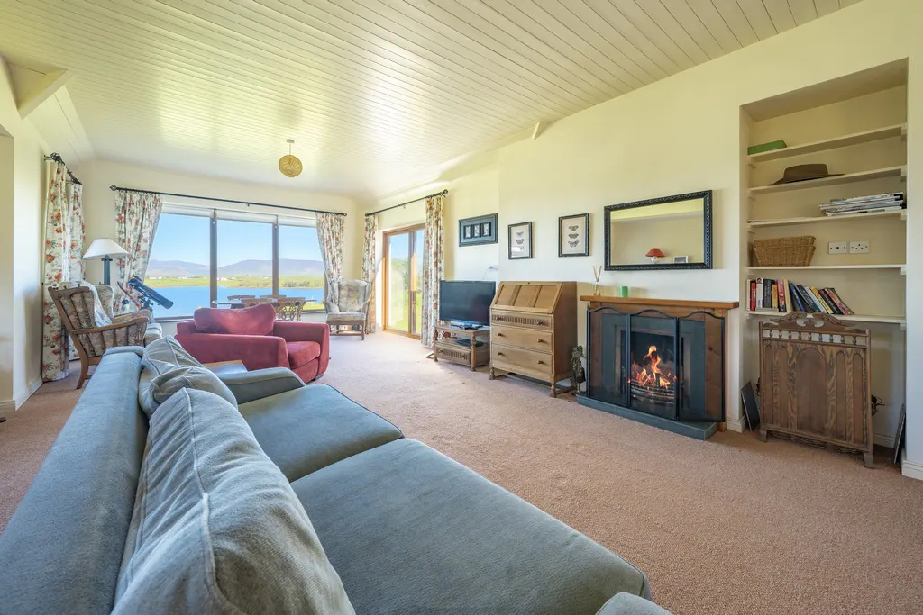 Living room with a gray sofa, red armchairs, and a fireplace with a burning fire. A large window overlooks a lake.