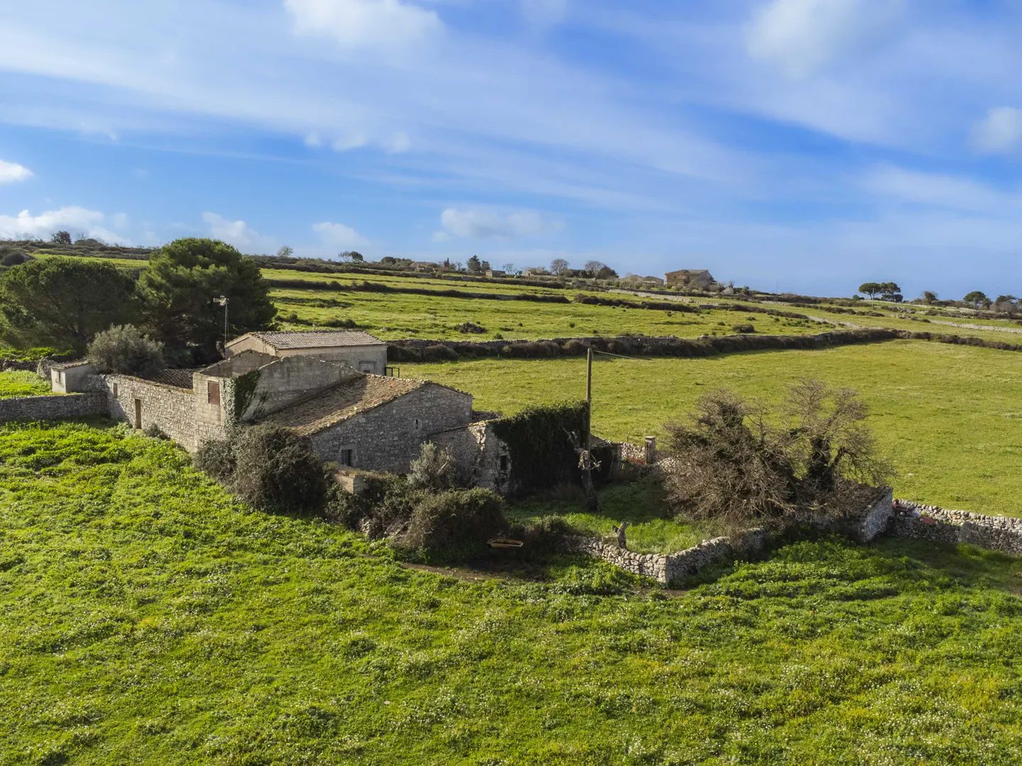Aerial view of a stone farmhouse with a tile roof, surrounded by green fields and stone walls under a blue sky.