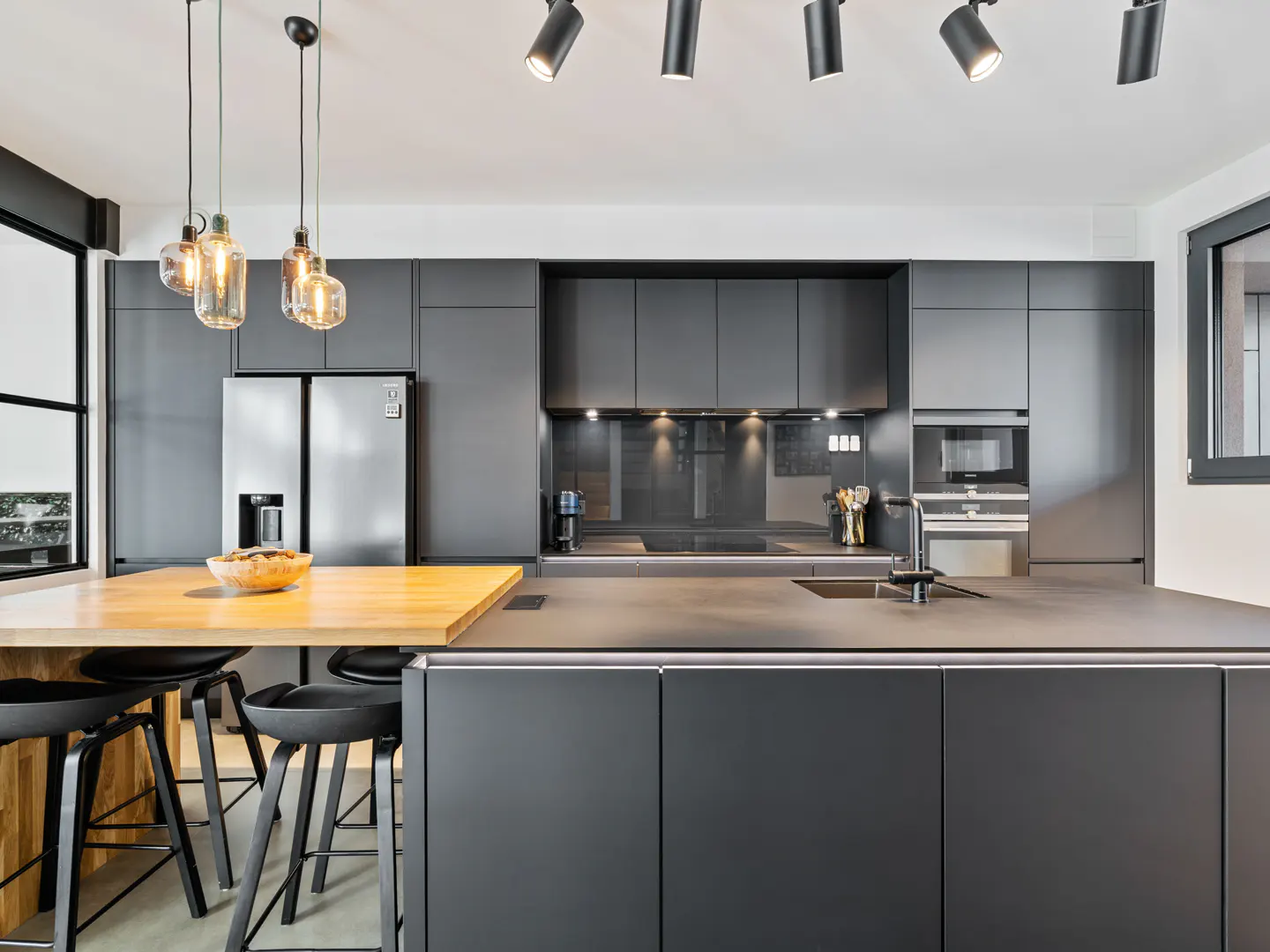 Modern kitchen with dark gray cabinets, stainless steel refrigerator, wood countertop island with black stools, and track lighting.