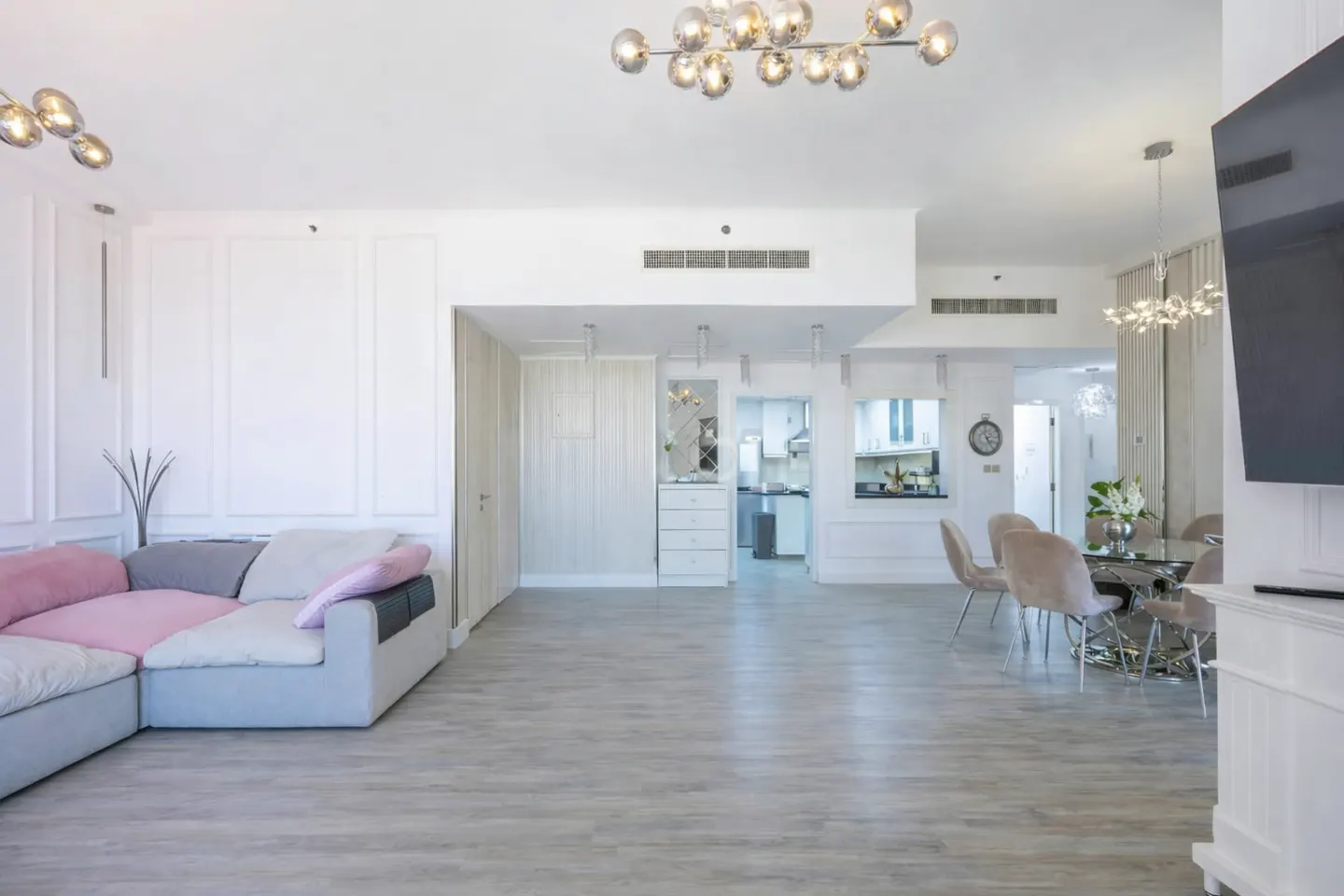 Bright, open-concept living space with a gray and pink sectional sofa, light wood floors, and a modern chandelier. Dining area and kitchen visible in the background.