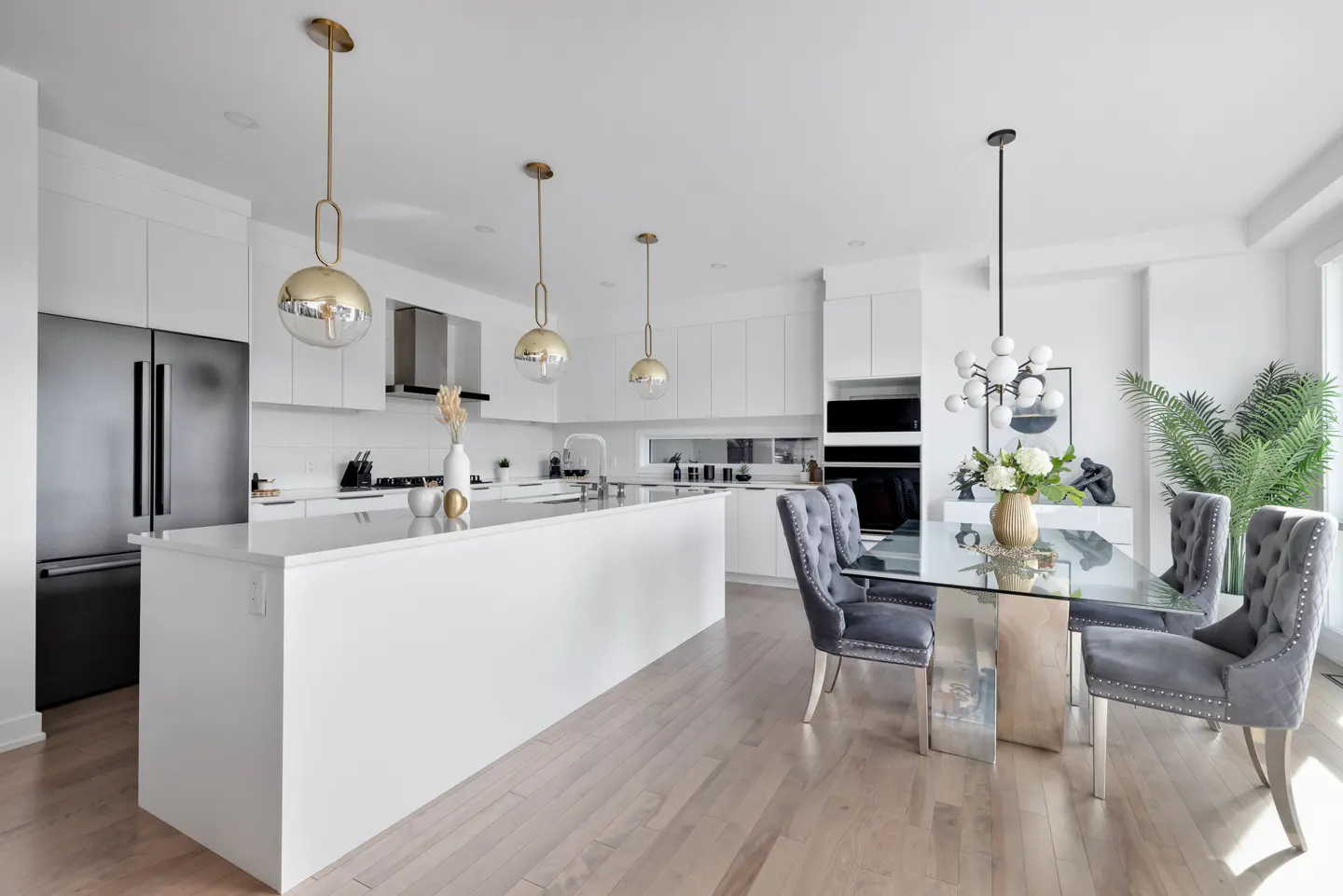 Bright, modern kitchen with white cabinets, stainless steel appliances, and a large island. A glass dining table with grey velvet chairs sits nearby.