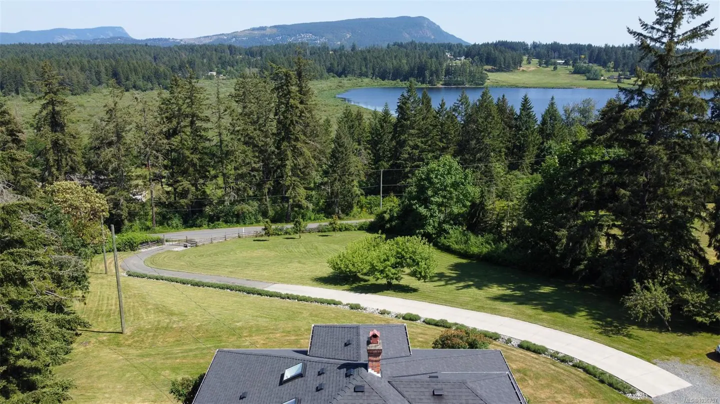 Aerial view of a house with a black roof, a long driveway, green grass, trees, a lake, and mountains in the background.
