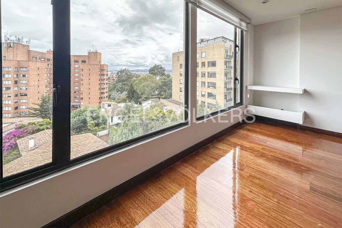 Empty room with polished hardwood floors, large windows, and a view of city buildings and trees under a cloudy sky.