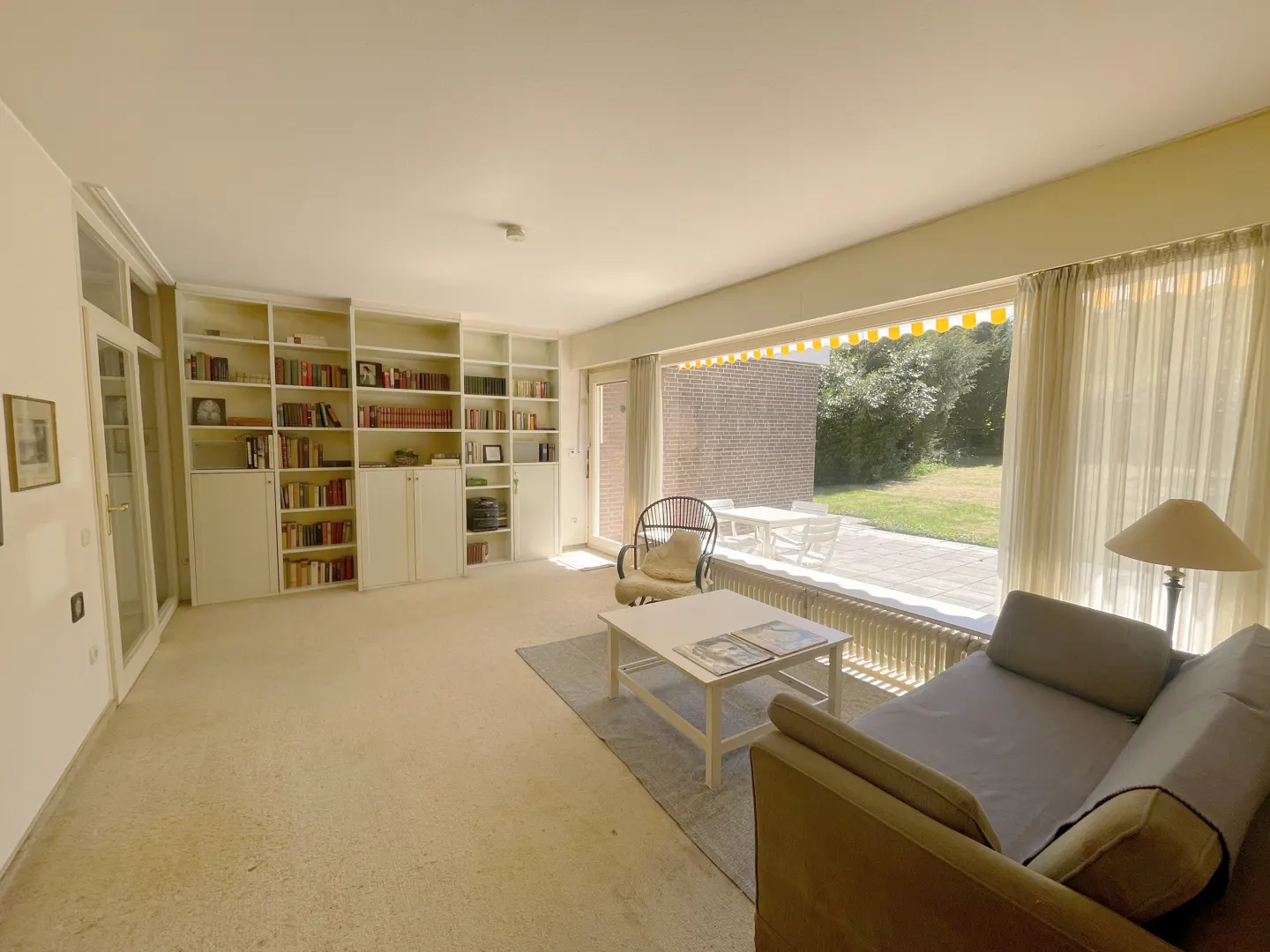 Bright living room with built-in white bookshelves, a gray sofa, and a large window overlooking a patio with white furniture.