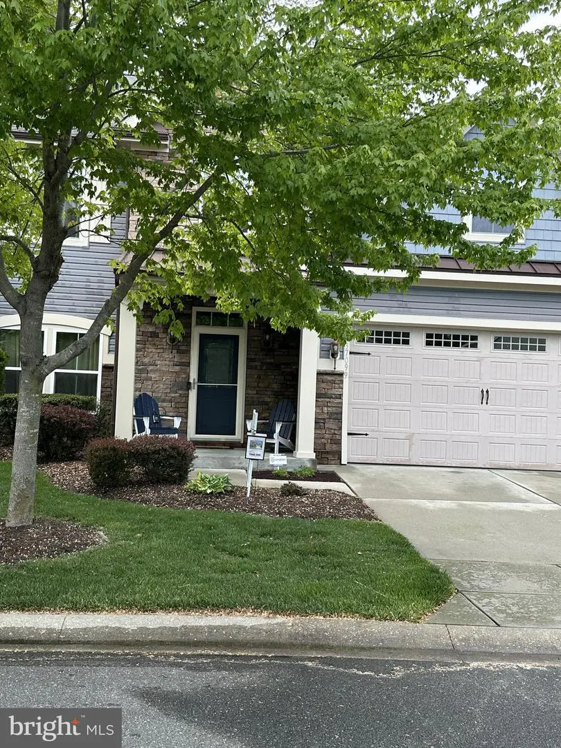 A two-story house with a stone facade, a white garage door, and a blue front door. Two blue chairs sit on the porch. A green tree shades the house.