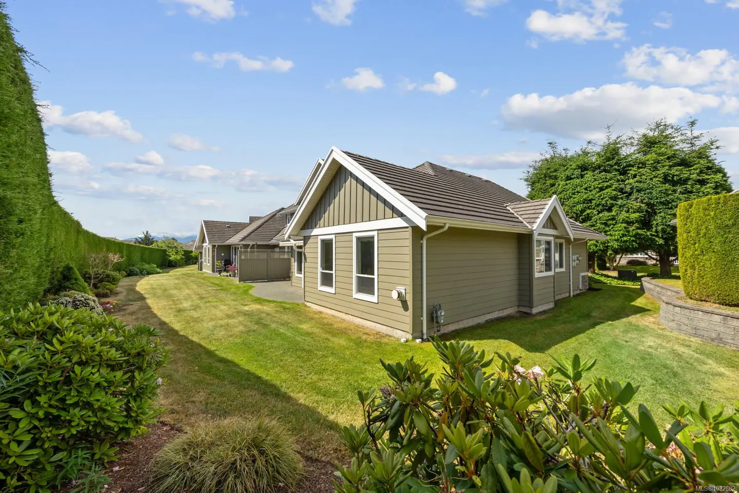 Exterior view of a sage green house with a dark roof, surrounded by a manicured lawn and tall hedges under a blue sky.
