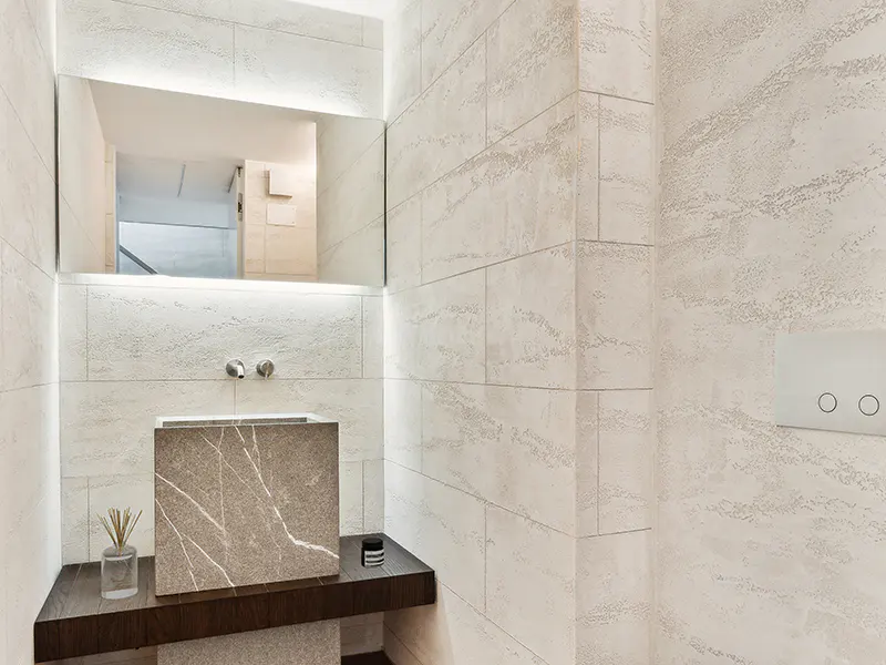 Modern bathroom with textured beige tile walls, a marble sink, and a dark wood shelf. A mirror with LED lighting is above the sink.