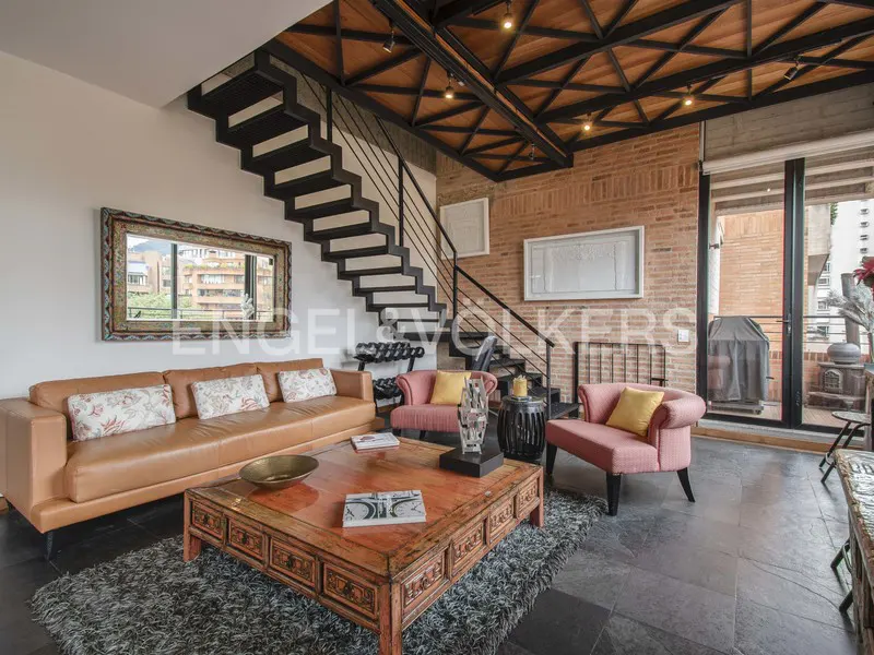 Living room with exposed brick wall, black staircase, and wood ceiling. A tan leather sofa and pink chairs sit on a gray rug.