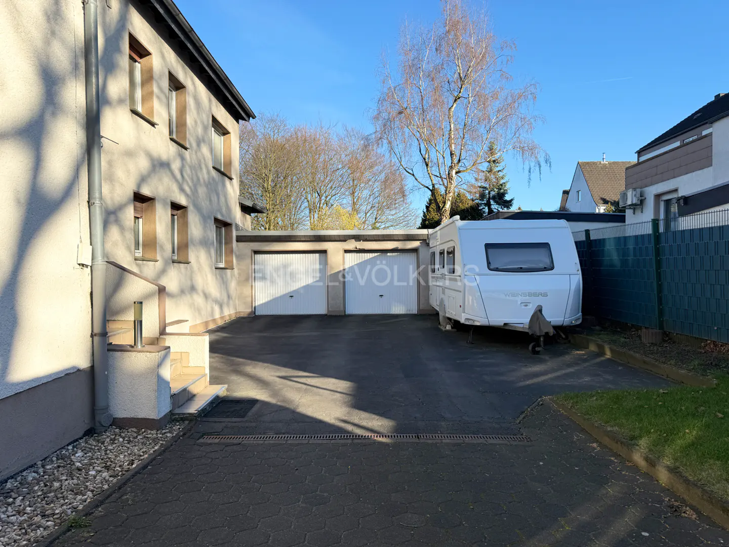 A paved driveway leads to a double garage next to a house, with a white caravan parked on the side under a clear blue sky.