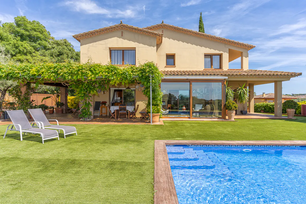 Two-story beige house with a blue tiled pool, green lawn, and lounge chairs. Vines cover a pergola near an outdoor dining area.